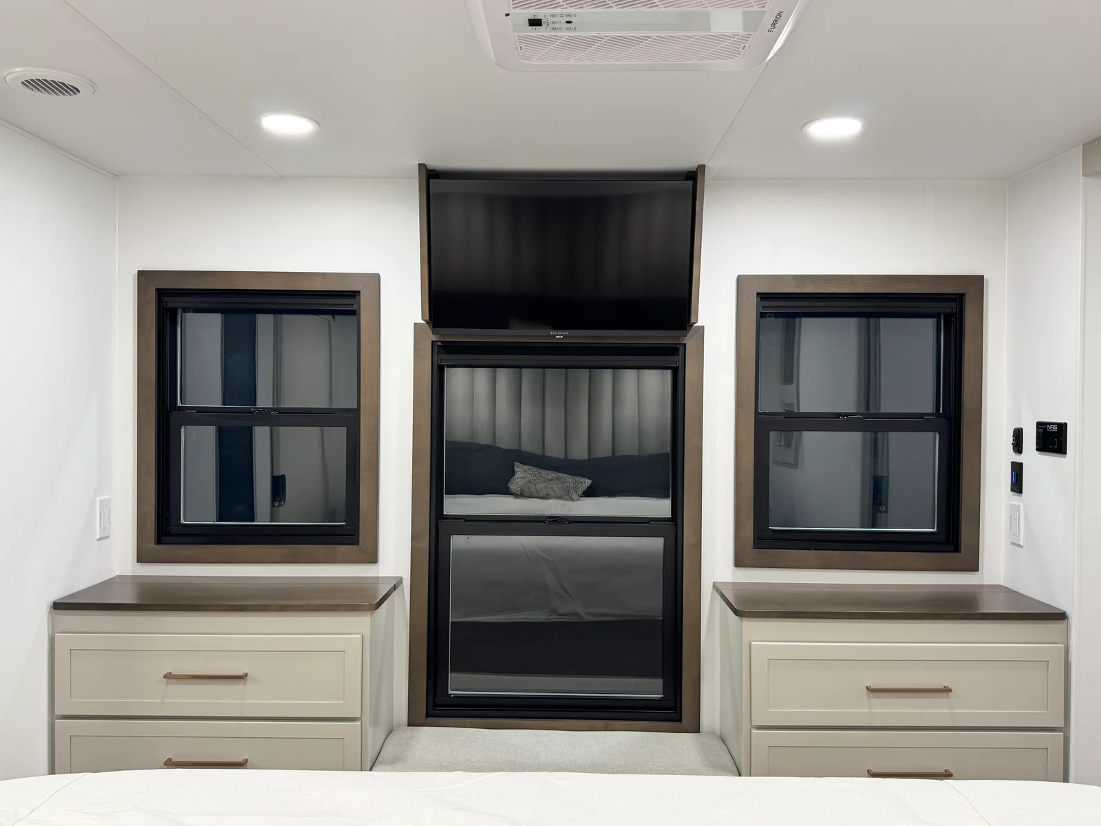 Foot of the Aurum bedroom with a wall-mounted TV above a window, flanked by two framed windows and dressers with warm metal pulls.