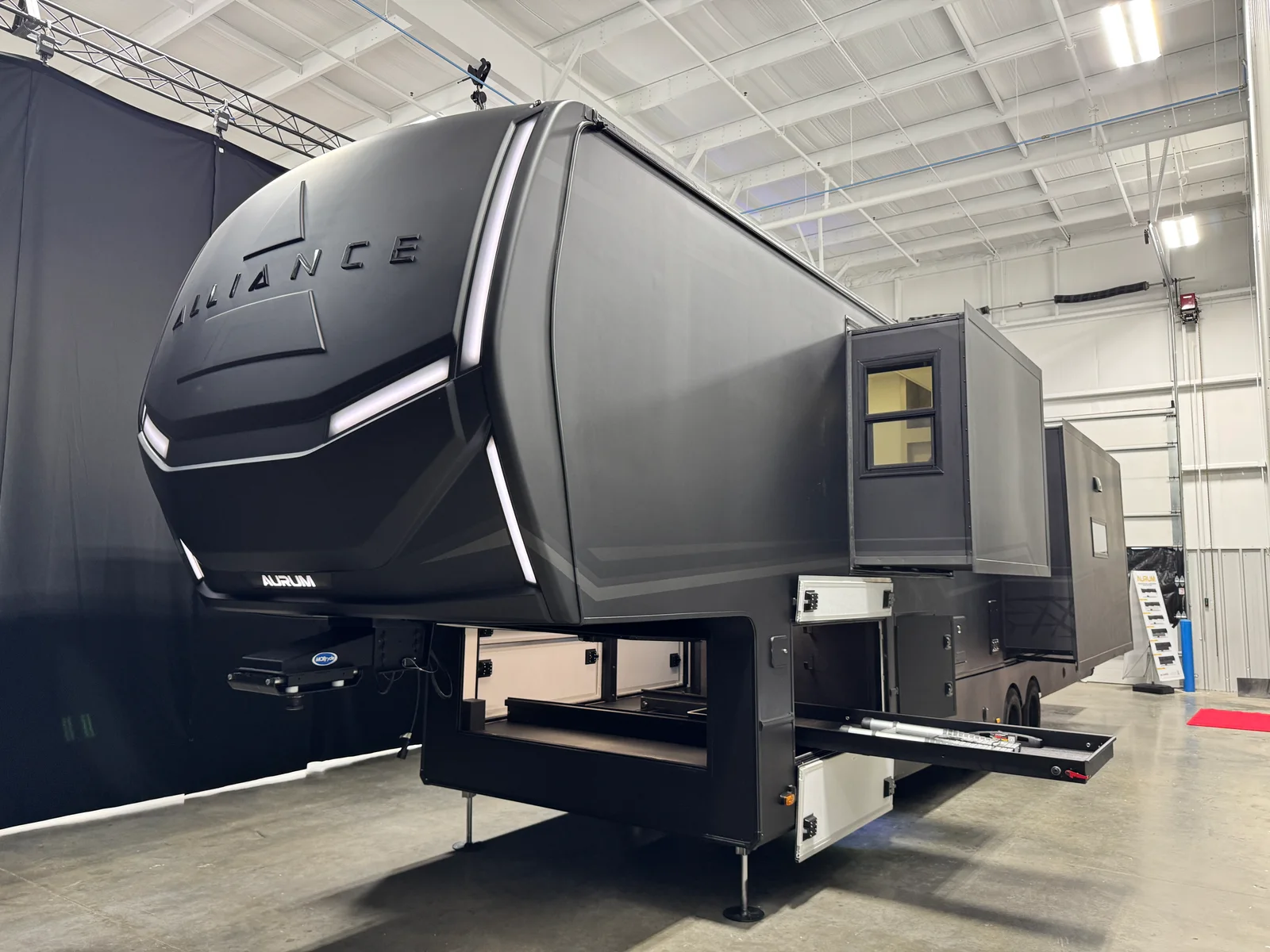Front three-quarter view of the dark Alliance Aurum with slideouts and exterior storage trays extended indoors.