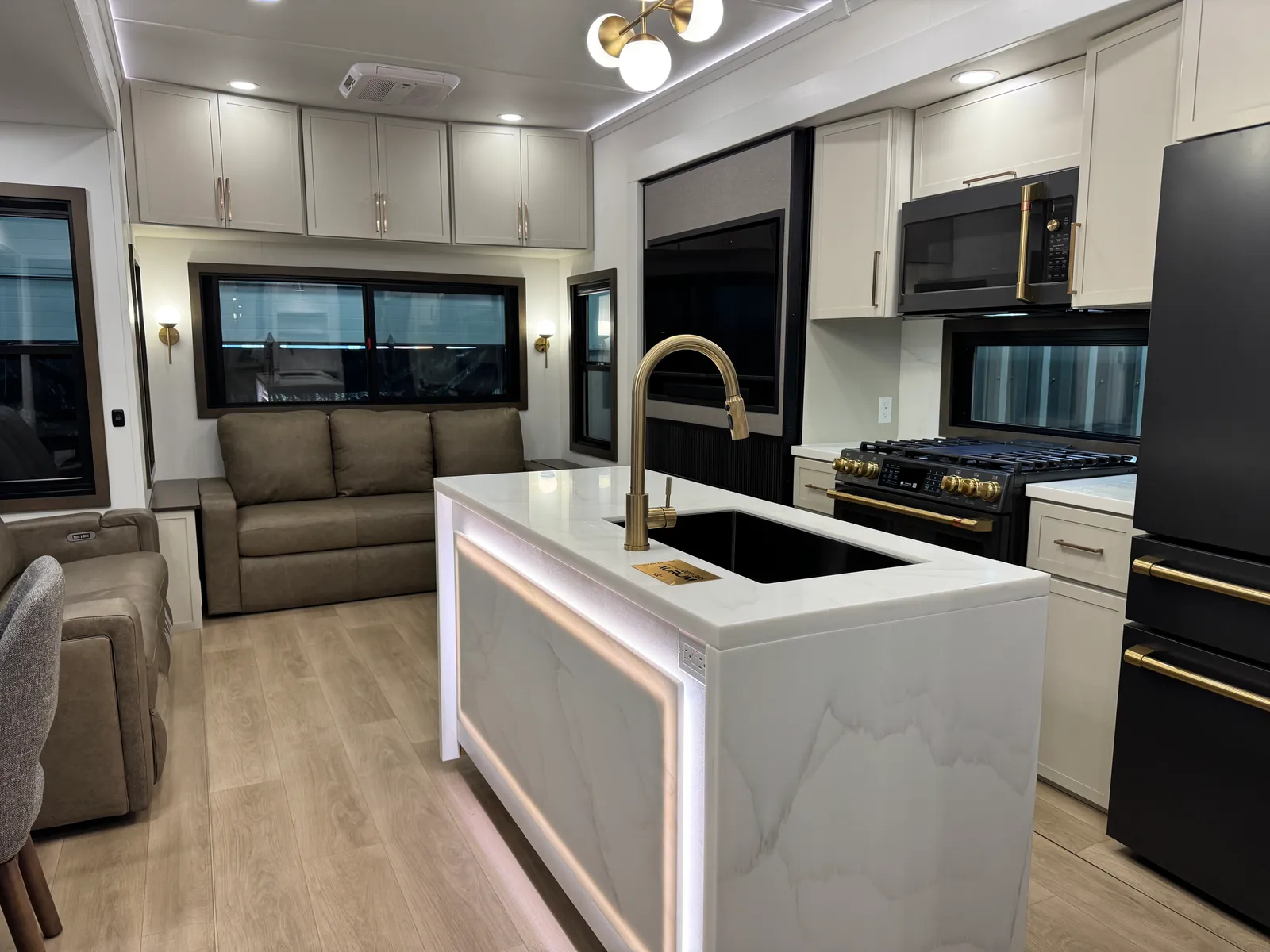 Kitchen island with a black undermount sink and gold faucet, lit underneath, beside the range and refrigerator.