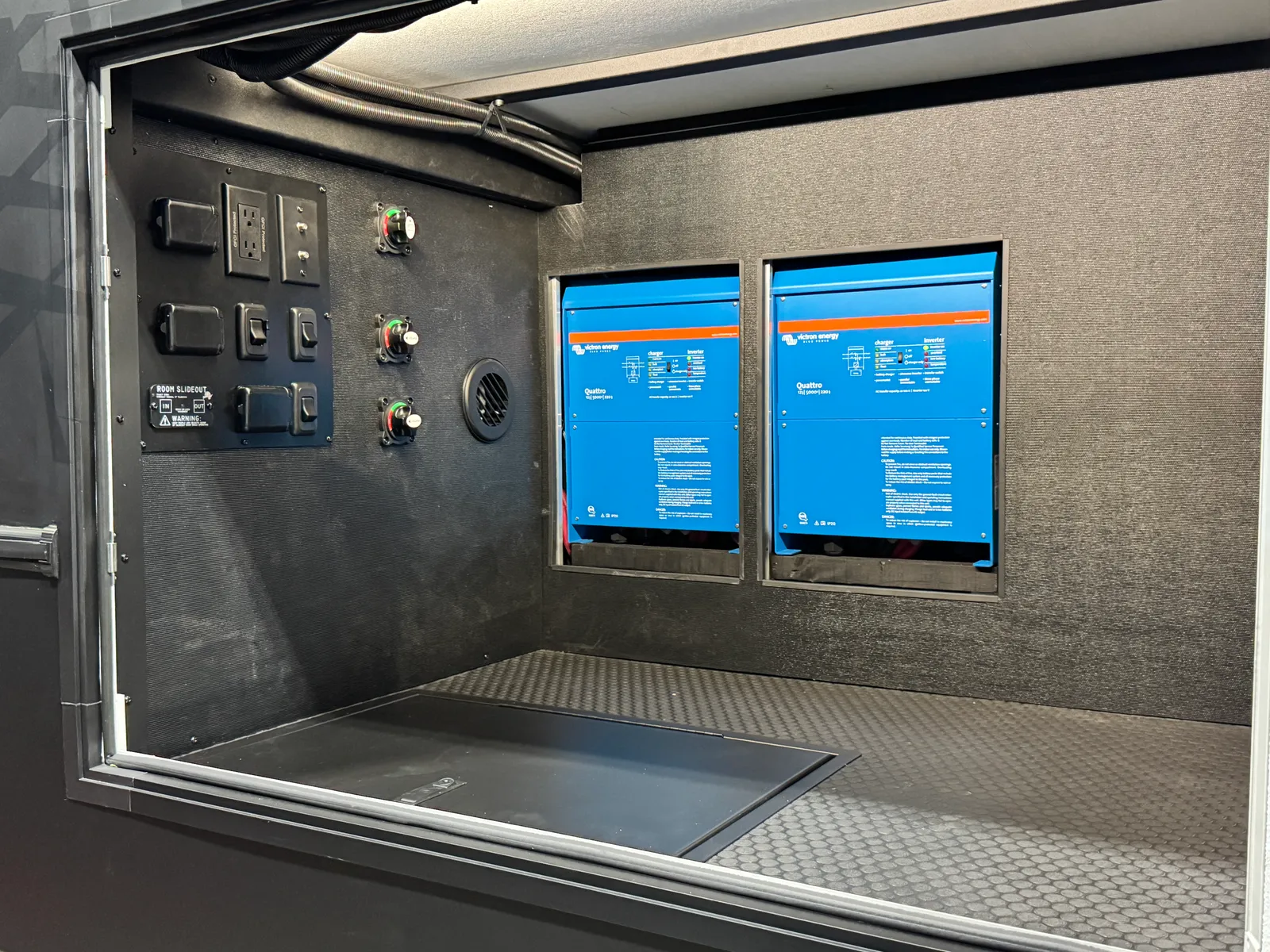 Two blue Victron Quattro inverters mounted in the lit storage bay beside a switch panel.